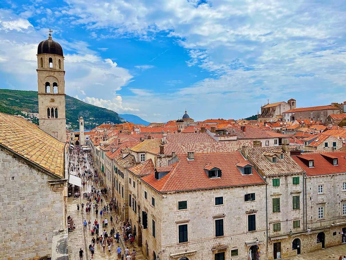 A bustling view of Stradun Street in Dubrovnik, with tourists walking between the old town's distinctive terracotta-roofed buildings and the prominent bell tower standing against a blue sky with wispy clouds.