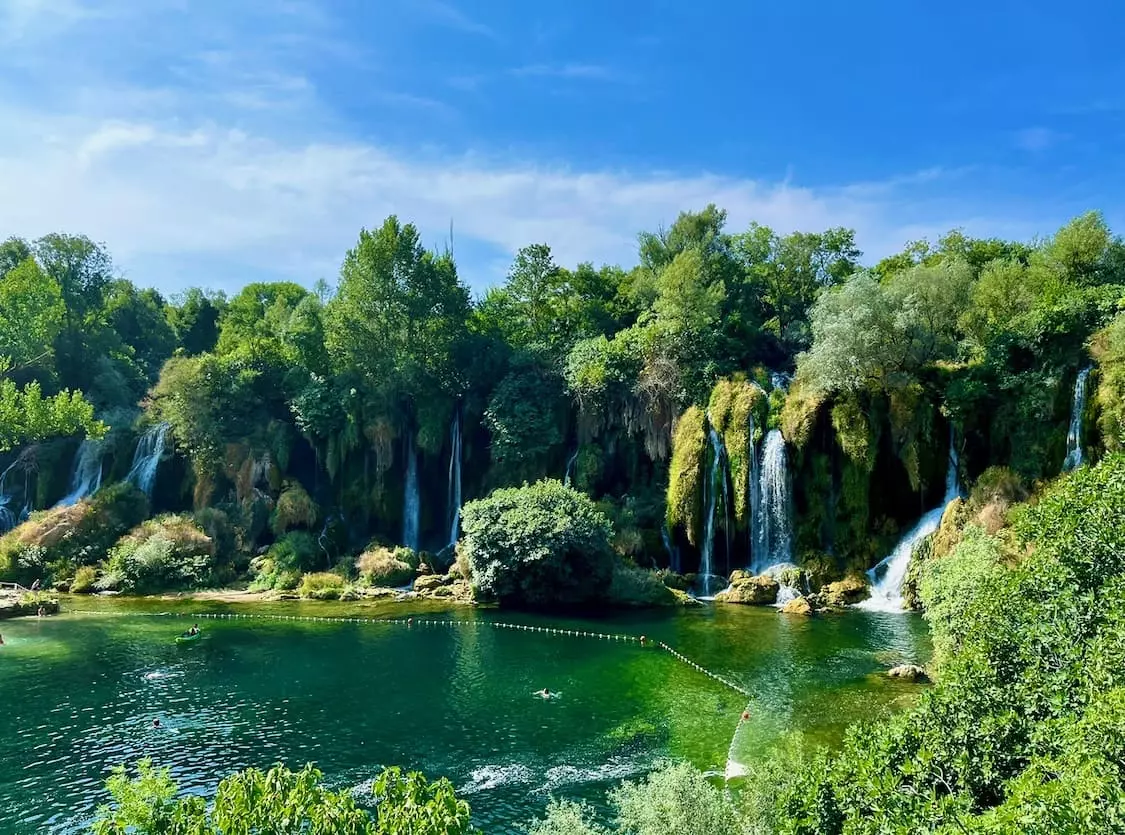 The serene Kravice Waterfalls in Bosnia, with its lush greenery and multiple cascades flowing into a emerald lake, viewed on a sunny day with a clear blue sky above.