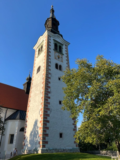 The Church of the Assumption of Mary on Bled Island, Slovenia. The tall white bell tower features red-brick accents, small arched windows, and a clock face near the top, set against a bright blue sky and surrounded by lush greenery.