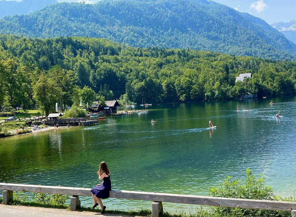 A woman in a blue dress sits on a wooden railing, gazing at the calm, emerald-green waters of Lake Bohinj in Slovenia. Paddleboarders and small boats are seen in the distance drifting across the lake, surrounded by dense forests and towering mountains under a clear blue sky. 