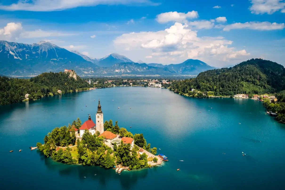 Aerial view of Lake Bled in Slovenia, showcasing the iconic Pilgrimage Church of the Assumption of Maria on an island, surrounded by emerald waters, with the Julian Alps and Bled Castle in the background on a sunny day