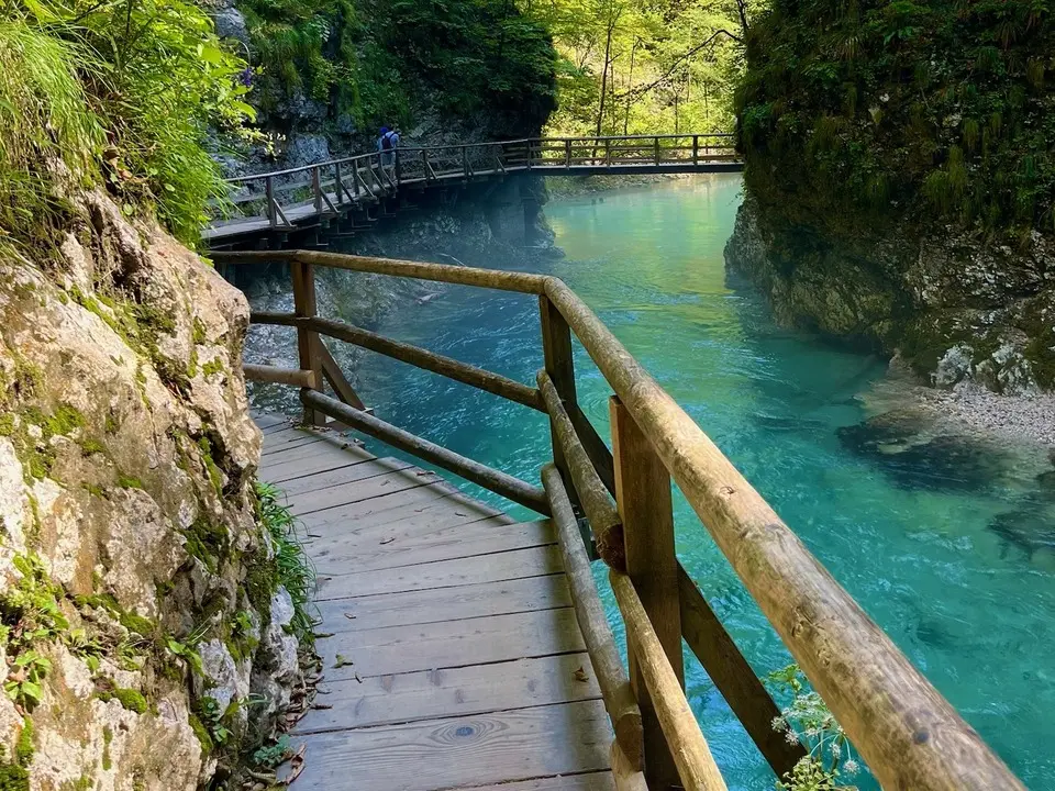 A wooden boardwalk in Vintgar Gorge, Slovenia, with a handrail winding along the edge of Radovna Valley, surrounded by rocky cliffs and green vegetation. A person can be seen in the distance walking along the path