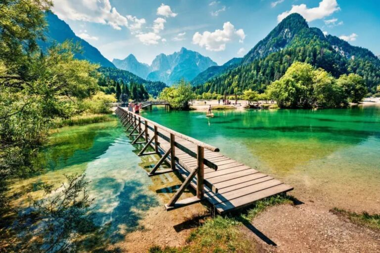 picturesque view of a lake in Slovenia, showcasing the country's natural beauty with a wooden jetty extending into the clear waters, framed by verdant trees and a majestic mountain range in the distance. Visitors enjoy the idyllic setting under a bright blue sky