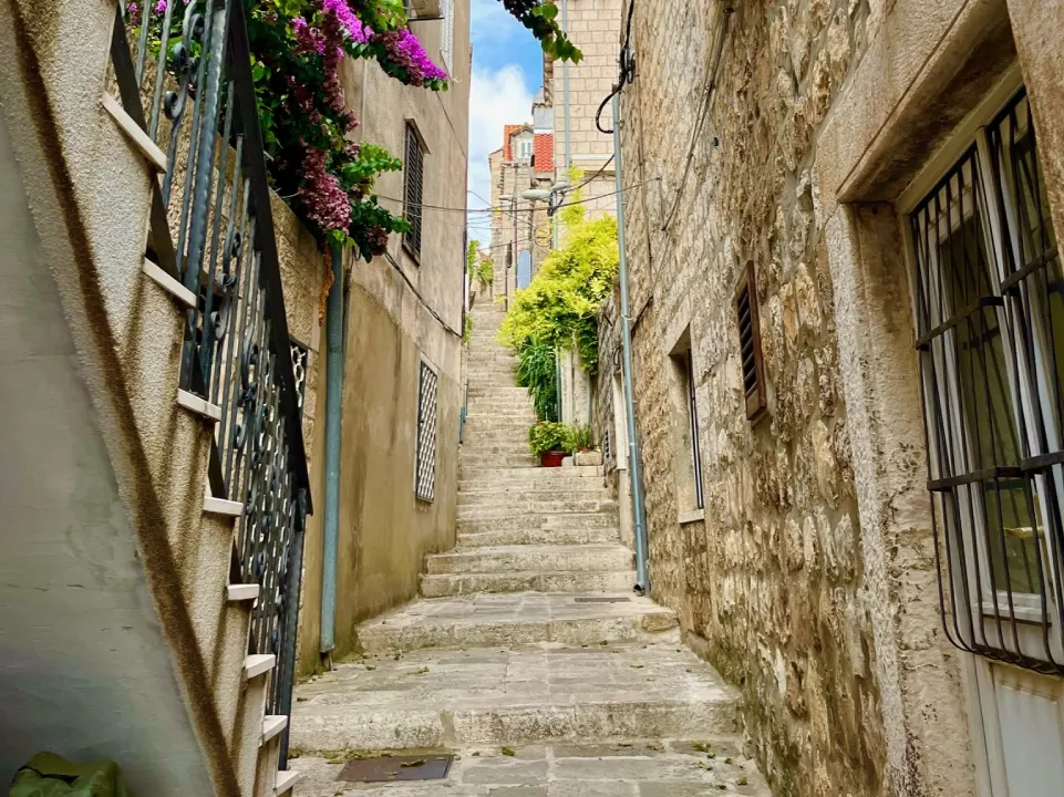 A narrow stone staircase in Cavtat, Croatia, between historic stone buildings. Vibrant purple flowers cascade over one wall. The bright blue sky peeks through above.