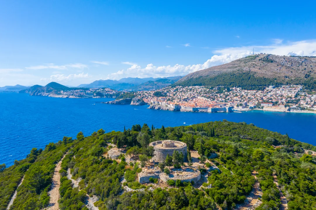 Aerial image showing Fort Royal on Lokrum Island, surrounded by greenery and set against the backdrop of the Adriatic Sea. In the distance, you can see the city of Dubrovnik and its iconic old town with terracotta rooftops.