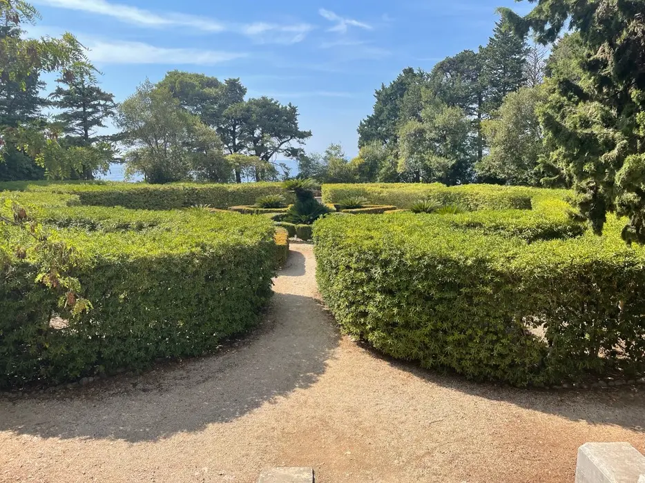 A sunny gravel path winding through the geometric hedge maze at the Benedictine Monastery of St. Mary on Lokrum Island, framed by lush trees with the Adriatic Sea in the background.