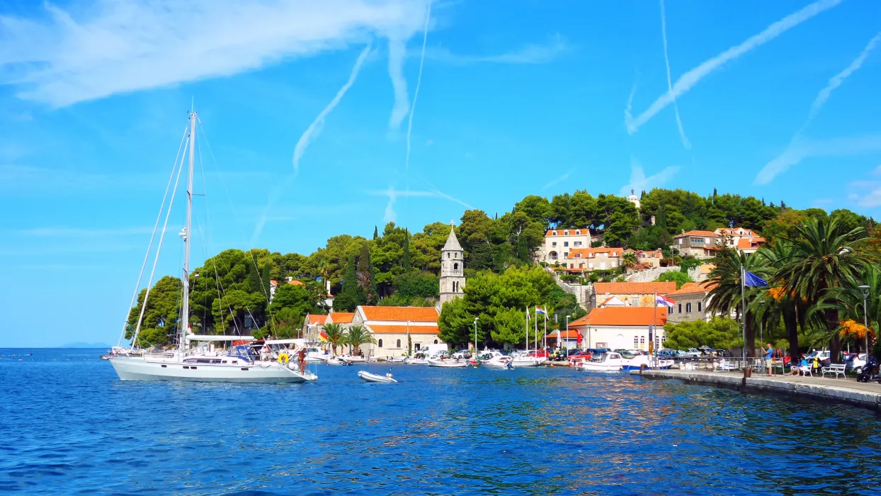 A sailboat cruising near the coastline of Cavtat, Croatia, with a backdrop of dense Mediterranean greenery, historical architecture, and a lively promenade under a vivid blue sky with wispy clouds