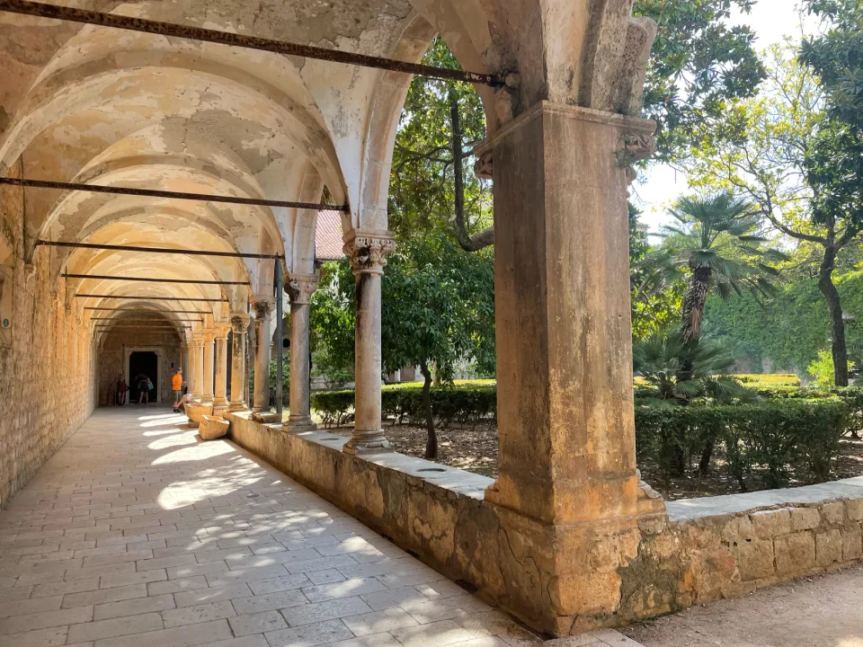 A sunlit stone cloister with archways and columns at the Benedictine Monastery on Lokrum Island. The walkway is lined with a low stone wall on one side, and greenery with palm trees in the garden to the right.