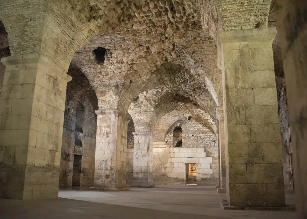 The cellars of Diocletian's Palace in Split, showcasing the architectural heritage with its sturdy Roman arches and columns. The vaulted stone ceilings and the serene ambiance hint at the palace's storied past, with a modest stone display in the foreground adding to the historic experience.