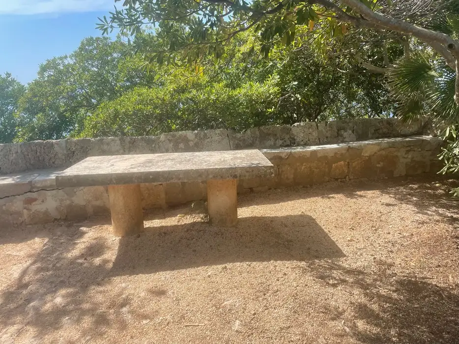 A weathered stone bench under the shade of leafy trees, situated on a gravel ground with a view of greenery in the background, on Lokrum Island.