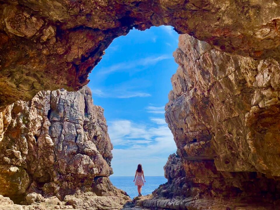 A woman in a white dress stands at the entrance of a rocky cove, looking at the bright blue sky and sea in the distance. The natural rock formations frame the view, creating a dramatic archway.