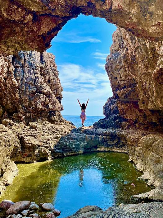 A woman holding her arms up at the edge of a rocky cove, framed by jagged cliffs and a natural arch, overlooking the Adriatic Sea
