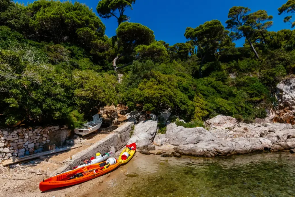 Two colourful kayaks rest on the shore amidst a backdrop of dense Mediterranean trees and rocky terrain on Lokrum Island, suggesting adventure in a tranquil cove