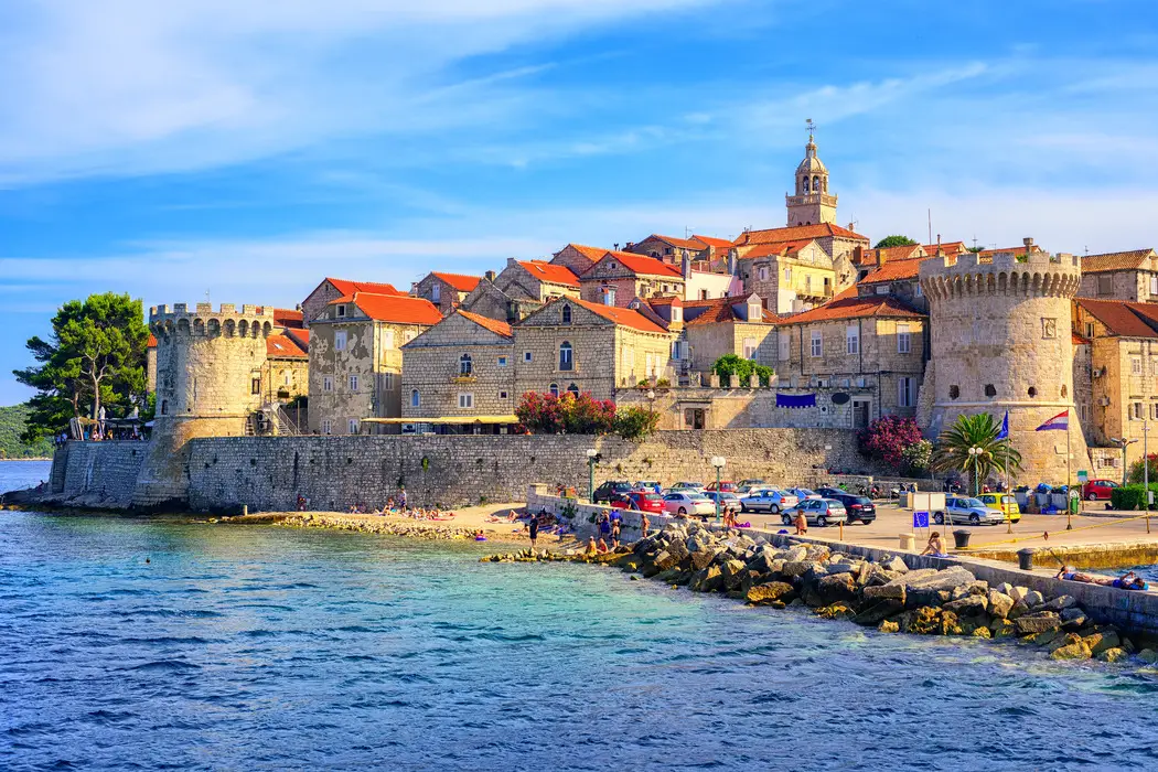The historic Korčula town with its medieval fortified walls and towers overlooking the Adriatic Sea, as people enjoy the nearby beach and vibrant streets under a clear sky in Croatia.