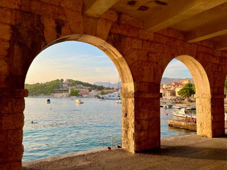 A set of stone archways framing a view of Cavtat's waterfront with boats and a coastal town in the background at sunset.