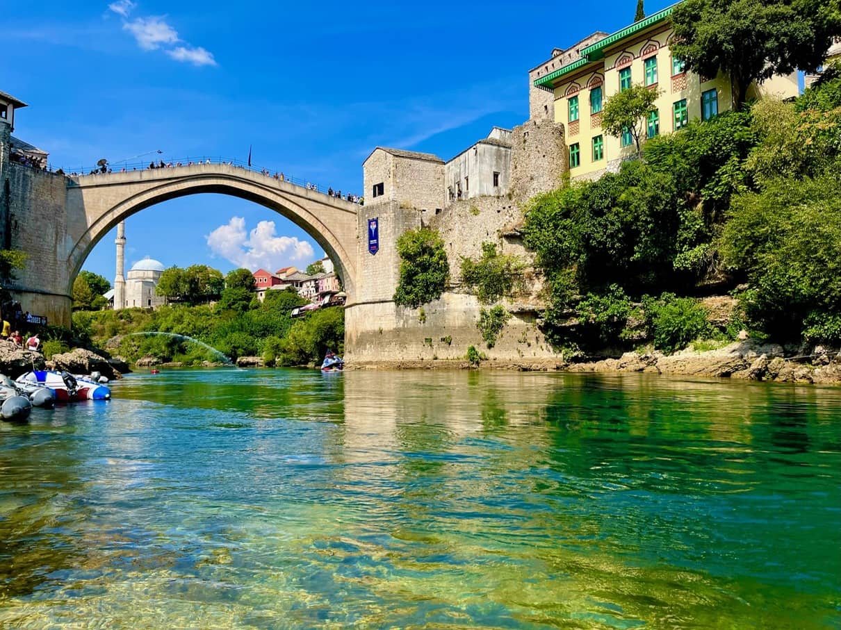 View of the iconic Stari Most (Old Bridge) in Mostar, Bosnia and Herzegovina, seen from the banks of the emerald-green Neretva River. The historic arch bridge connects two medieval towers against a backdrop of bright blue skies, while visitors enjoy boating activities on the river.