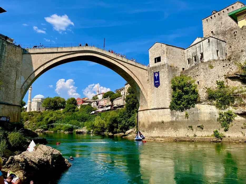 The Stari Most bridge arching over the emerald waters of the Neretva River in Mostar, Bosnia and Herzegovina, with spectators lining the railing and historic city buildings in the background under a clear blue sky