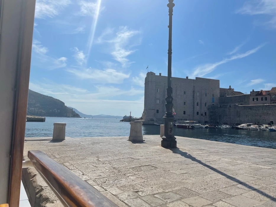 View of the Old Port of Dubrovnik with historic fortifications overlooking the calm Adriatic Sea, under a sunny sky with a light post casting a shadow on the stone pavement