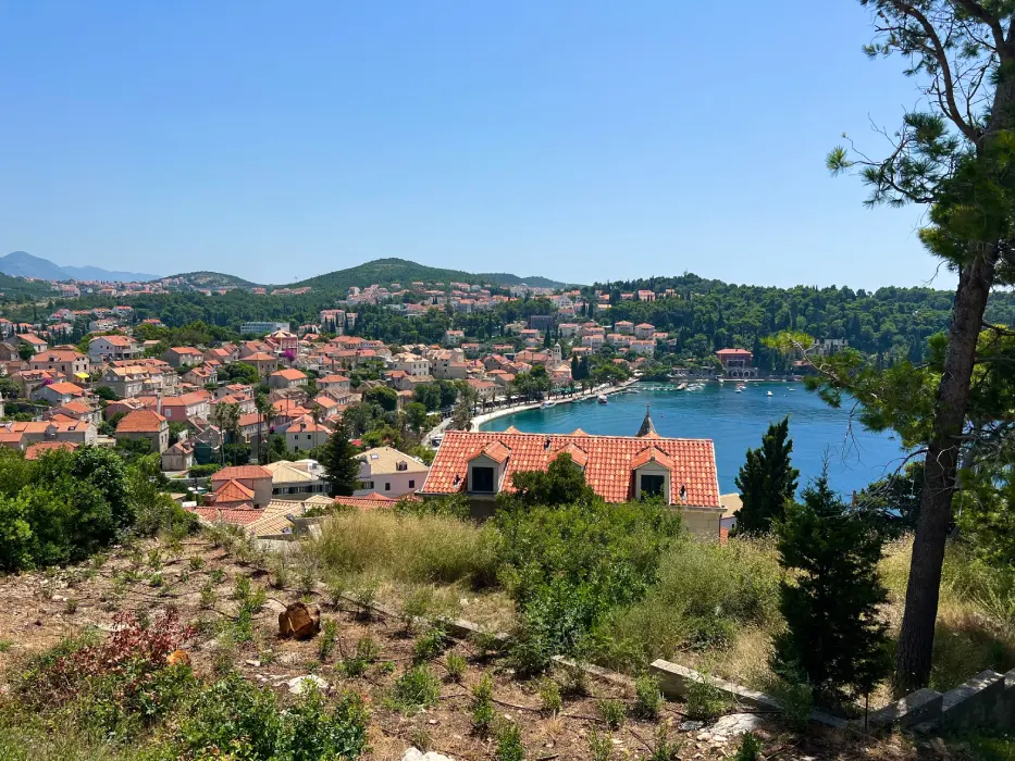 Panoramic view overlooking the terracotta rooftops of Cavtat, Croatia, with the serene blue waters of the Adriatic Sea and lush green hills in the background, under a clear sky.