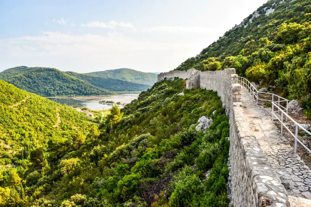 Ancient stone city walls of a ston, Croatia, winding through lush green hillsides, overlooking a serene lake in a valley, under a clear sky.