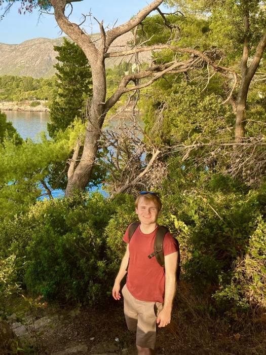 Person smiling on a sunny day on the Sustjepan Hiking Trail in Cavtat, surrounded by trees with a blue lake and mountainous landscape in the distance.