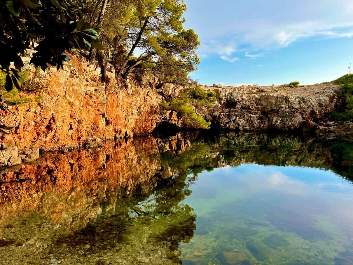 The Dead Sea Lake on Lokrum Island by reddish cliffs and green trees, with water reflecting the vibrant colours of the cliffs and a clear blue sky above.