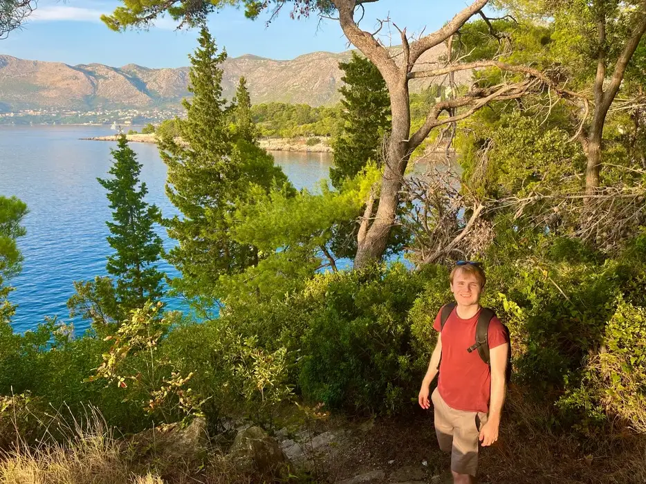 A person with a content smile, wearing a red shirt and khaki shorts, enjoys a sunny day on the Sustjepan Hiking Trail in Cavtat, surrounded by verdant trees with a tranquil blue lake and mountainous landscape in the distance.