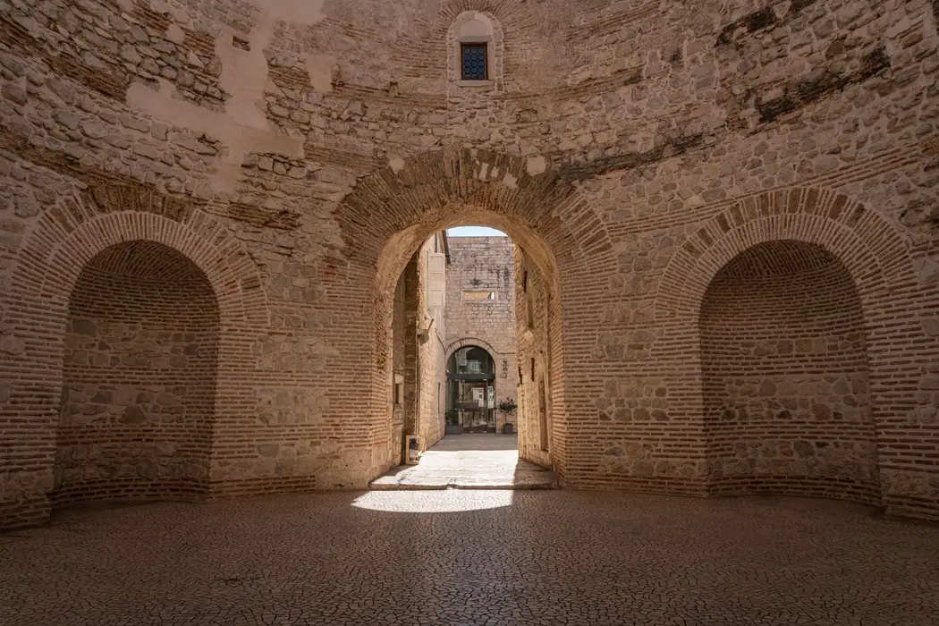Interior view of Diocletian Palaces historical stone archway leading to a small courtyard, with cobblestone pavement and a clear blue sky peeking through. The ancient walls show varying textures and patterns of brick and stone, creating a sense of old-world charm