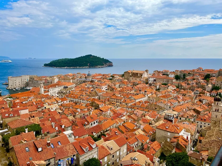 Panoramic view from the Dubrovnik city walls, showcasing the dense terracotta rooftops, historic architecture, and the Adriatic Sea with Lokrum Island in the distance.