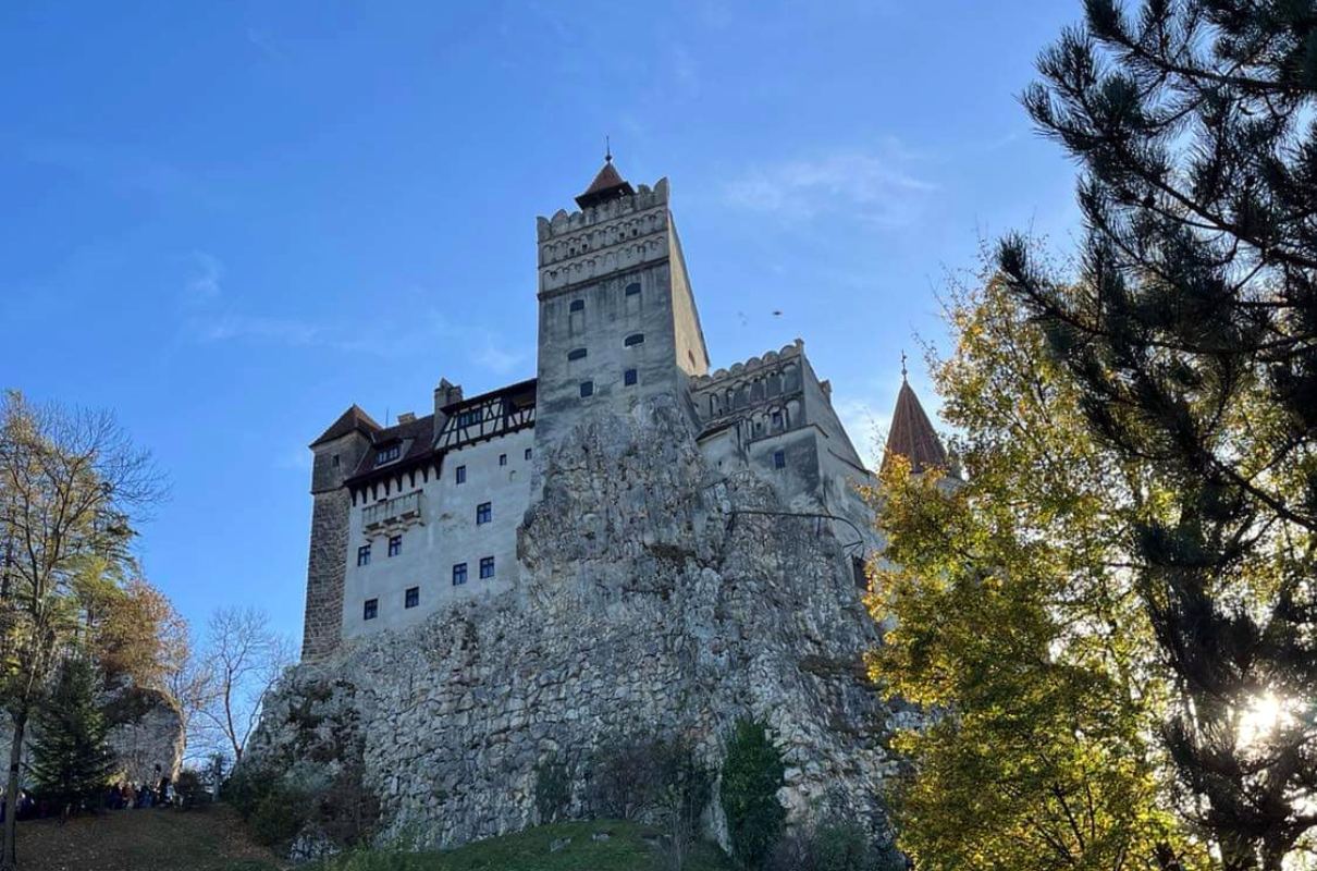 A view of Bran Castle perched atop a rocky outcrop, framed by autumn foliage and a clear blue sky, with sunlight filtering through the trees in the foreground.