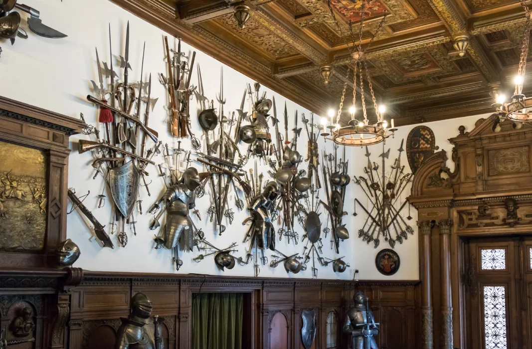 Historic armory display inside Peles Castle, Romania, showcasing a diverse collection of medieval weaponry and armor on a white wall, accented by rich wooden paneling and elegant chandeliers, evoking a sense of regal history and craftsmanship.