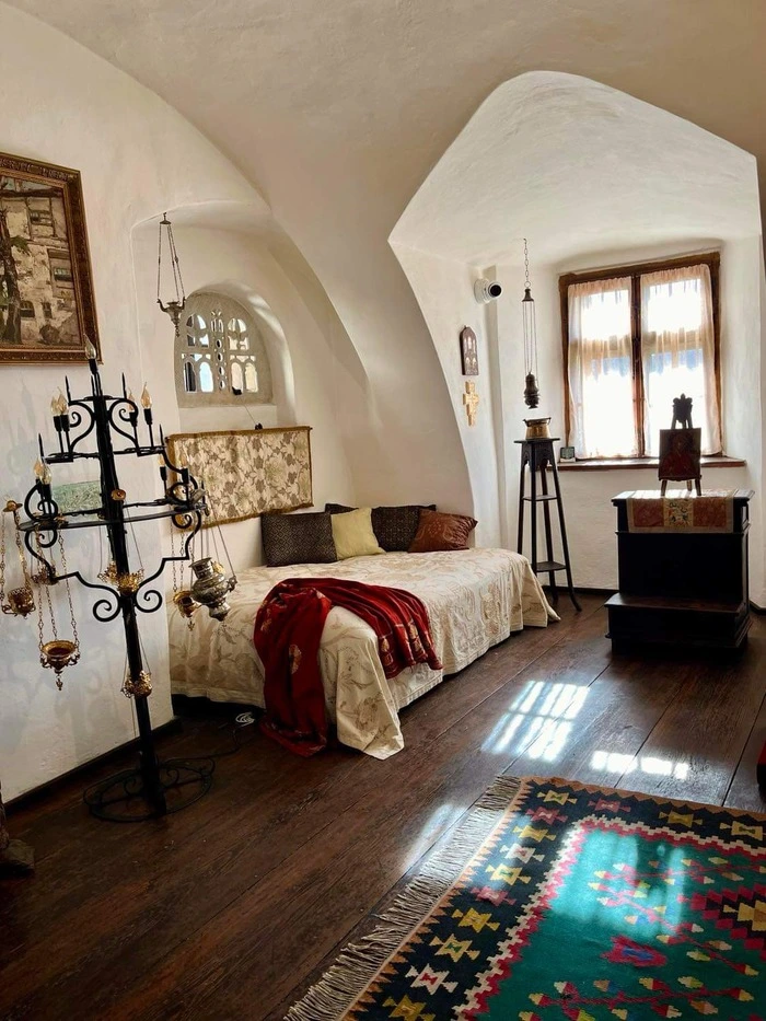 Vintage-style bedroom in Bran Castle, showcasing arched white ceilings, a candelabra, a bed with ornate patterns and a red throw, and a Southwestern rug, bathed in the natural light from a traditional window