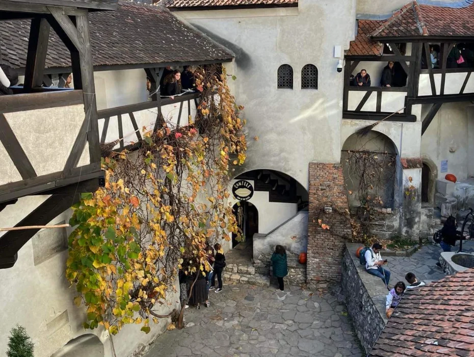 A bustling courtyard within Bran Castle with tourists exploring its medieval architecture, including a stone tower and timber balconies adorned with autumn leaves.