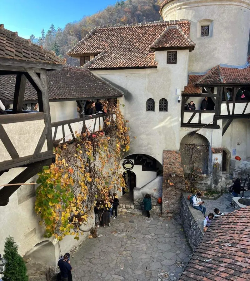 A bustling courtyard within Bran Castle with tourists exploring its medieval architecture, including a stone tower and timber balconies adorned with autumn leaves.