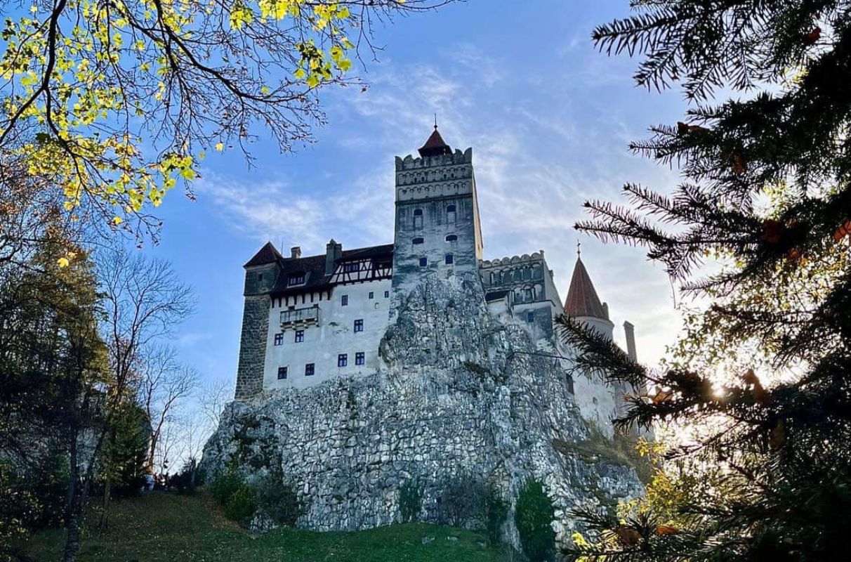 A view of Bran Castle perched atop a rocky outcrop, framed by autumn foliage and a clear blue sky, with sunlight filtering through the trees in the foreground.