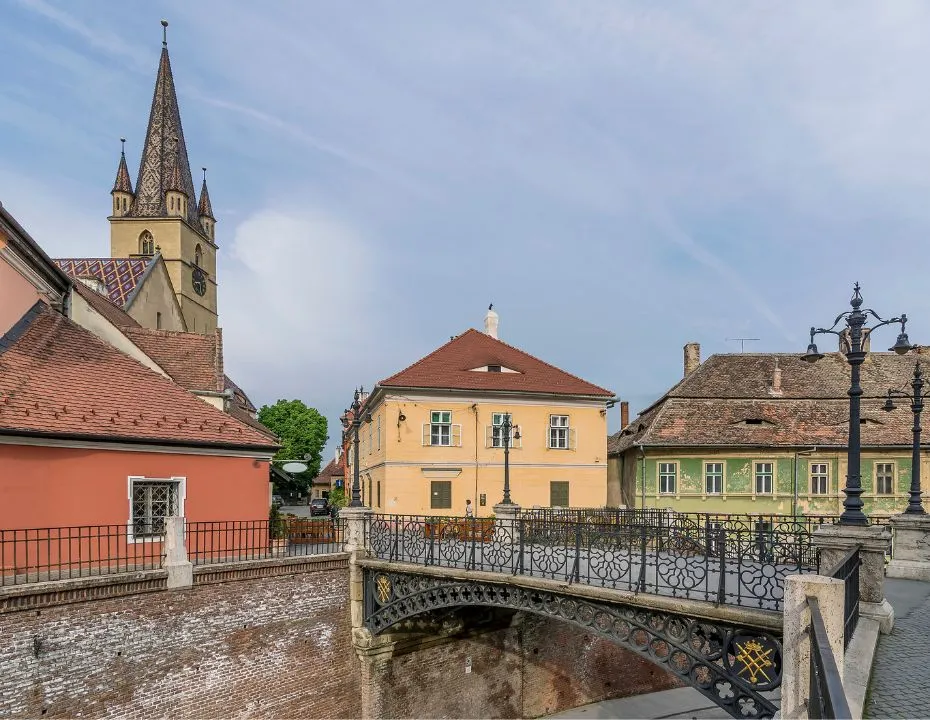 Scenic view during a day in Sibiu, featuring the iconic Bridge of Lies with its decorative railings, flanked by traditional vibrant buildings and the pointed steeple of a church piercing the blue sky.
