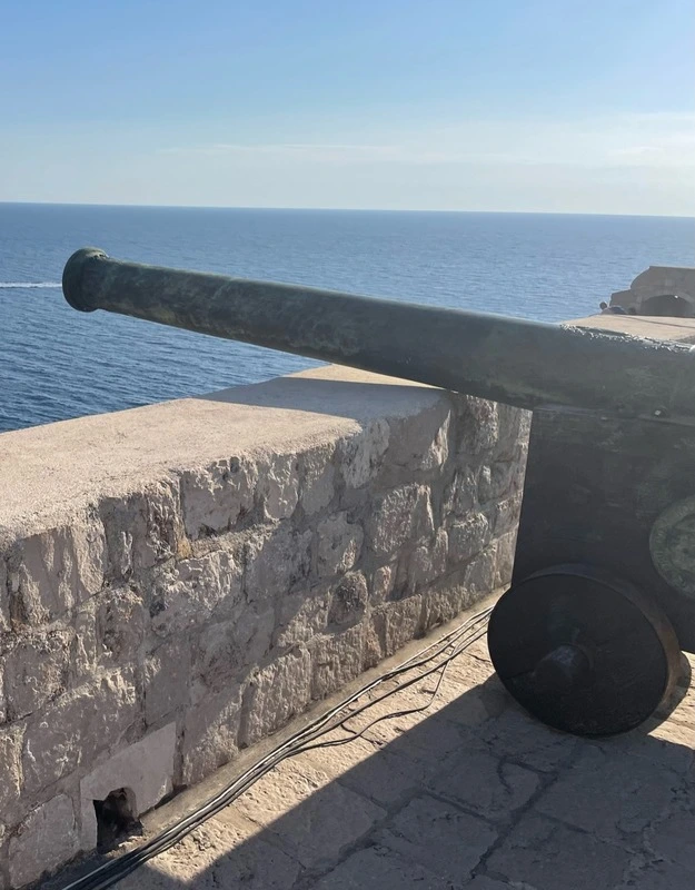 An old cannon perched atop the stone walls of Fort Lovrijenac in Dubrovnik, overlooking the Adriatic Sea under a clear sky