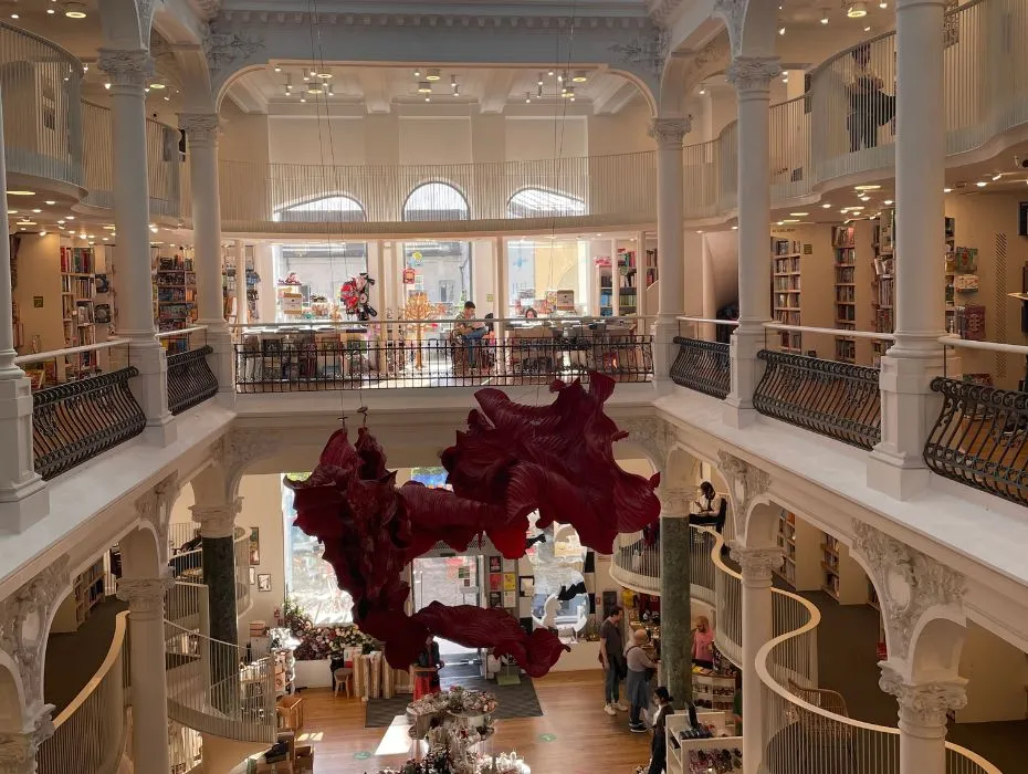 Interior view of Cărturești Carusel bookstore in Bucharest, showcasing a multi-level open space with ornate white balustrades, bookshelves filled with books, visitors browsing, and a unique red sculpture hanging in the centre