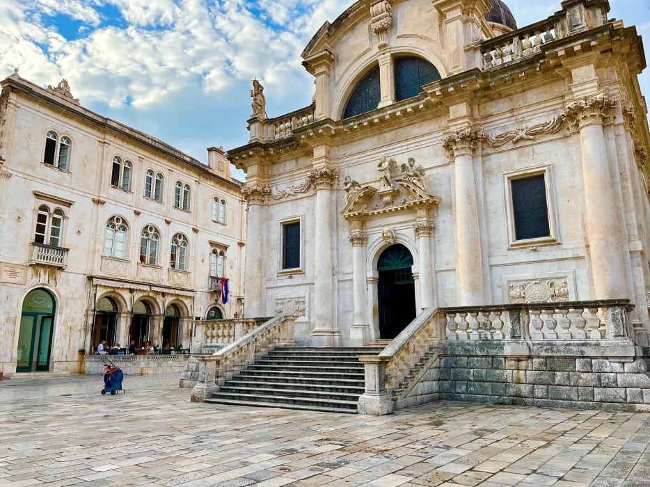 Exterior of the Church of St. Blaise in Dubrovnik, displaying Baroque architecture with ornate stone carvings and statues, under a partly cloudy sky. A person is crouching in the square, capturing the moment.