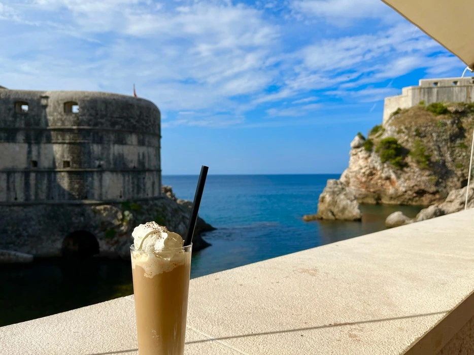 Iced coffee with whipped cream in the foreground, overlooking the circular Fort Bokar and the Adriatic Sea from the terrace of Dubravka 1836 restaurant & café in Dubrovnik, under a clear blue sky.