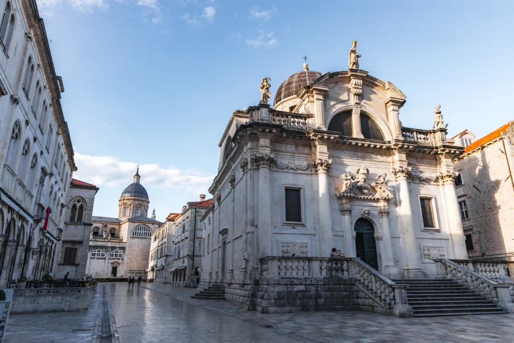 Dubrovnik Cathedral with its central dome and baroque façade, by stone buildings on a quiet, sunlit street.