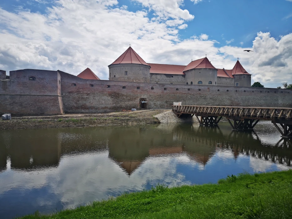 Medieval fortress in Romania with a wooden bridge reflected in the moat under a cloudy sky.