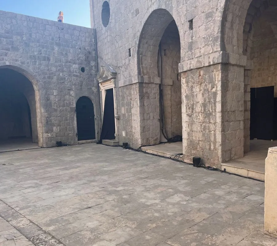 Sunlit courtyard of Fort Lovrijenac in Dubrovnik with robust stone walls and arches, where a lone person stands on the upper battlements overlooking the area.