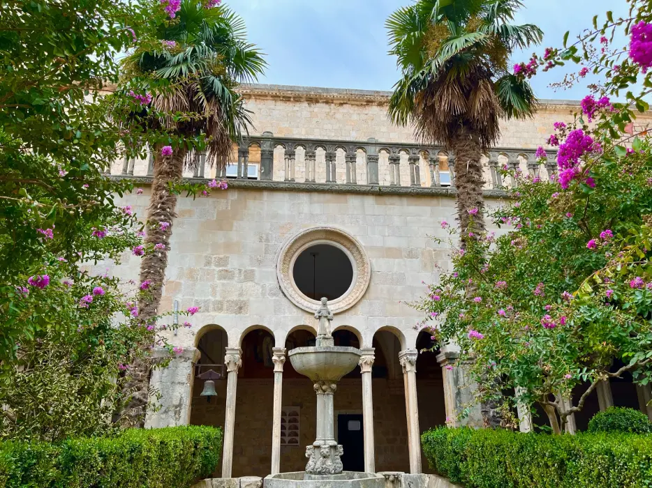 Courtyard of the Franciscan Monastery in Dubrovnik, featuring a central fountain, surrounding columns, vibrant bougainvillea, and palm trees under a partly cloudy sky.
