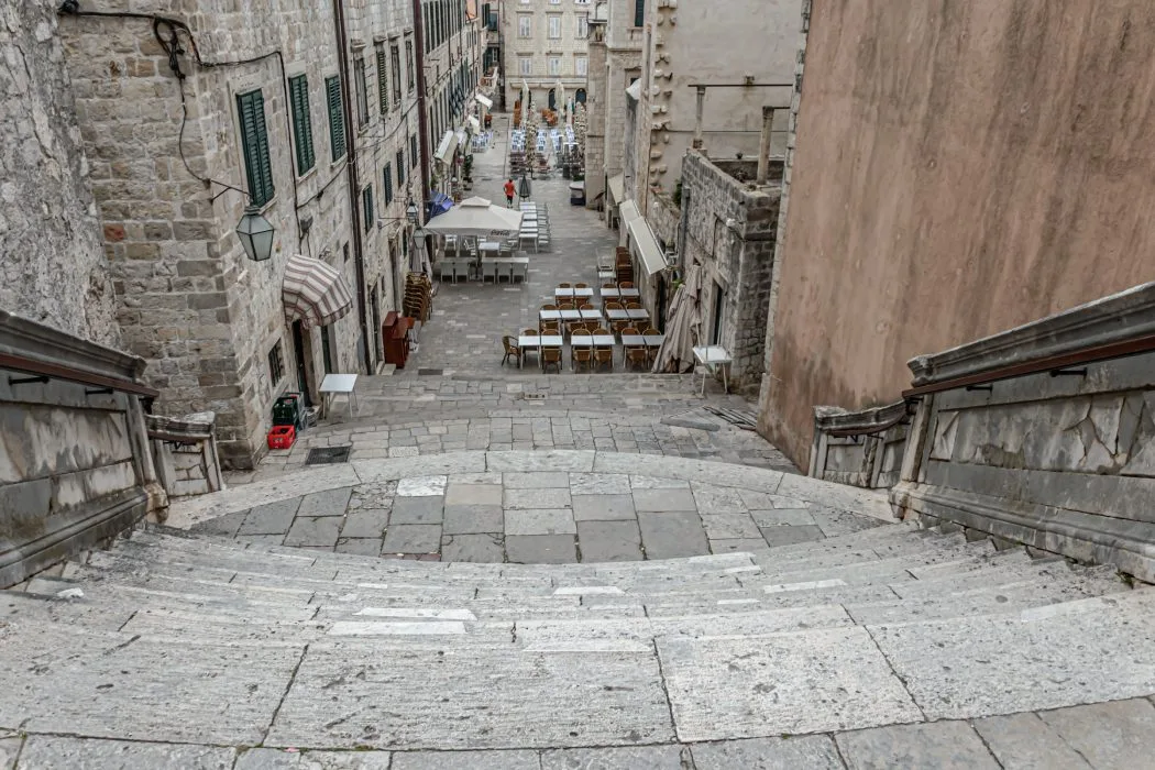 View from the top of the iconic Jesuit Stairs in Dubrovnik, often recognized as a filming location for the 'Game of Thrones' series, leading down to a historic street lined with stone buildings and outdoor cafe seating