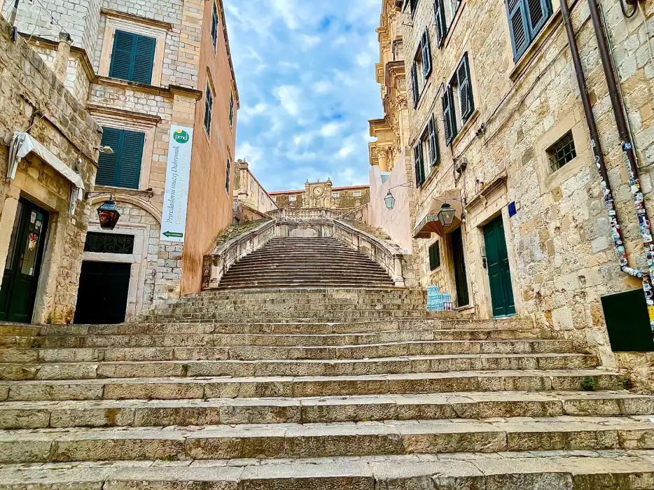The iconic Jesuit Staircase in Dubrovnik, known as a filming location for the "Game of Thrones" series, leading up to a stately building with Baroque architecture under a partly cloudy sky. Stone buildings with wooden shutters flank the staircase, adding to the historical charm of this popular tourist spot.
