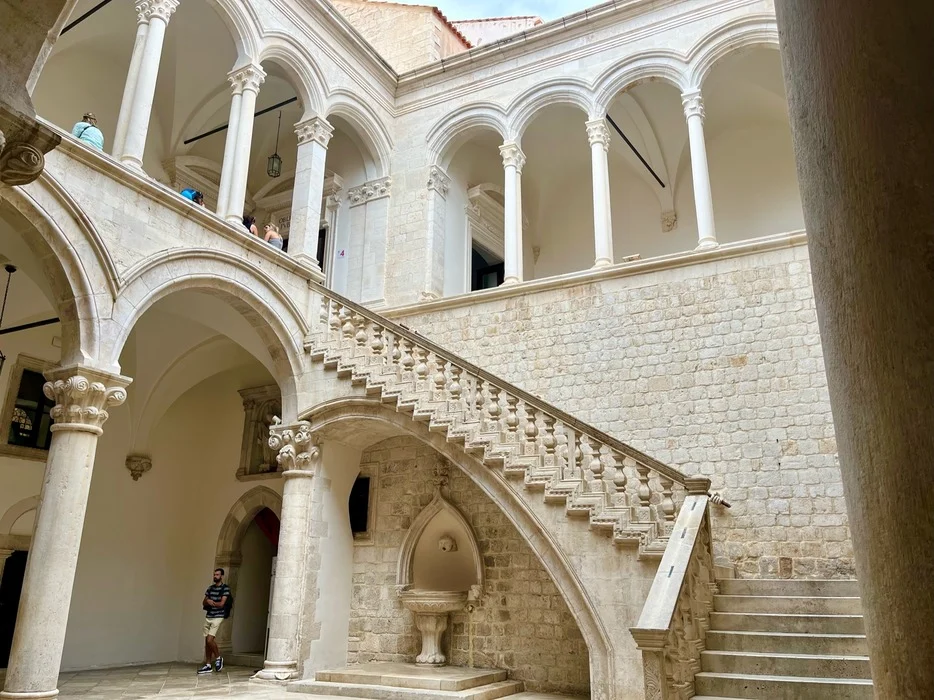 Elegant stone staircase and arched walkways within the Rector's Palace in Dubrovnik, known as a filming location for 'Game of Thrones', with visitors exploring the historical site.