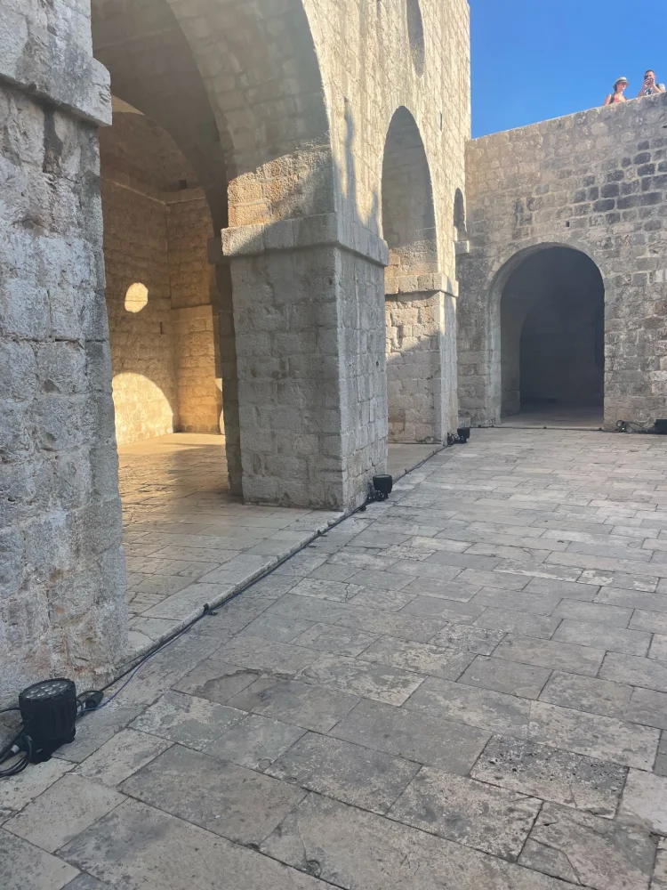 Inside Fort Lovrijenac in Dubrovnik, a sunlit courtyard showcases the robust stone architecture with arched passageways. A few visitors can be glimpsed in the distance on the fort's upper level, absorbing the ambiance of this 'Game of Thrones' filming site.