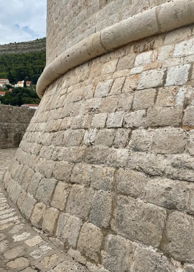 Curved stone walls of the historic Minčeta Tower in Dubrovnik, with a glimpse of the old city's terracotta roofs and lush hillside in the background, capturing the essence of Croatian heritage and a popular Game of Thrones filming location.