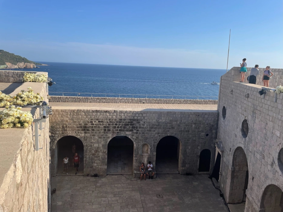 Visitors explore Fort Lovrijenac, a historic fortress with stone walls overlooking the Adriatic Sea in Dubrovnik, known for being a filming location for 'Game of Thrones'. People are seen standing on the fort's upper ramparts and in the shaded archways, enjoying the scenic ocean view and architecture."